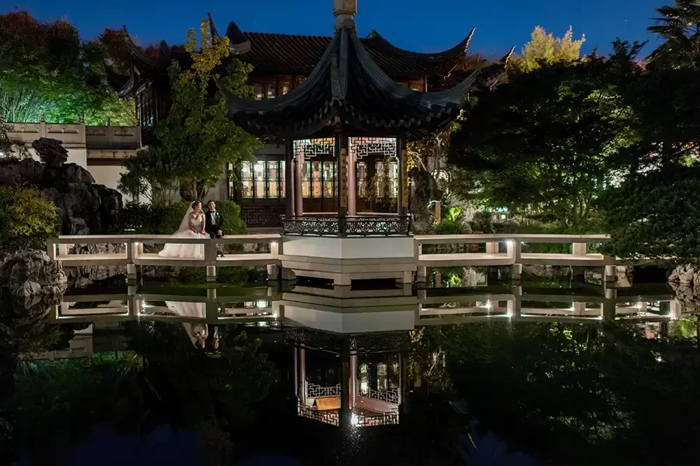 Portland Wedding Photographer Robert Knapp catches this photo with no flash. A photo across a water feature at the Chinese garden. The couple and the bridge they are standing on reflect in the water. 