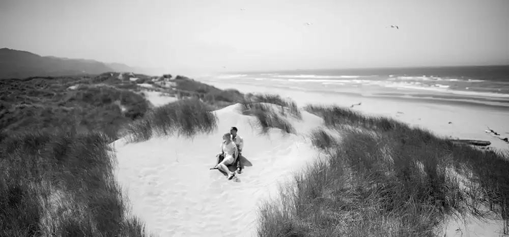 a couple sit in a sand dune with the ocean behind them a seagull flies over Cannon Beach Engagement Photos With Photographer Robert Knapp