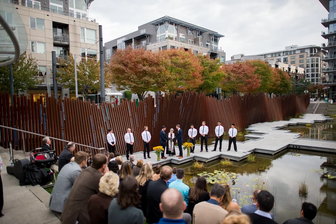the ceremony in the center of the park taking place on a dock over a water feature with sunflowers as decor, all of the wedding party has white button down shirts LGBT Wedding Photographer Robert Knapp at ​Tanner Springs Park in Portland Oregon