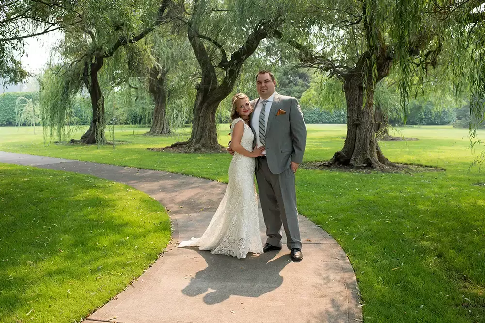 ​McMenamins Grand Lodge Weddings 
from Robert Knapp Photographer a bride and groom stand at the bend of a curved path. The sun shines down onto their shoulders from behind. The wedding dress of lace is spread across the path. Willow trees scatter across the background