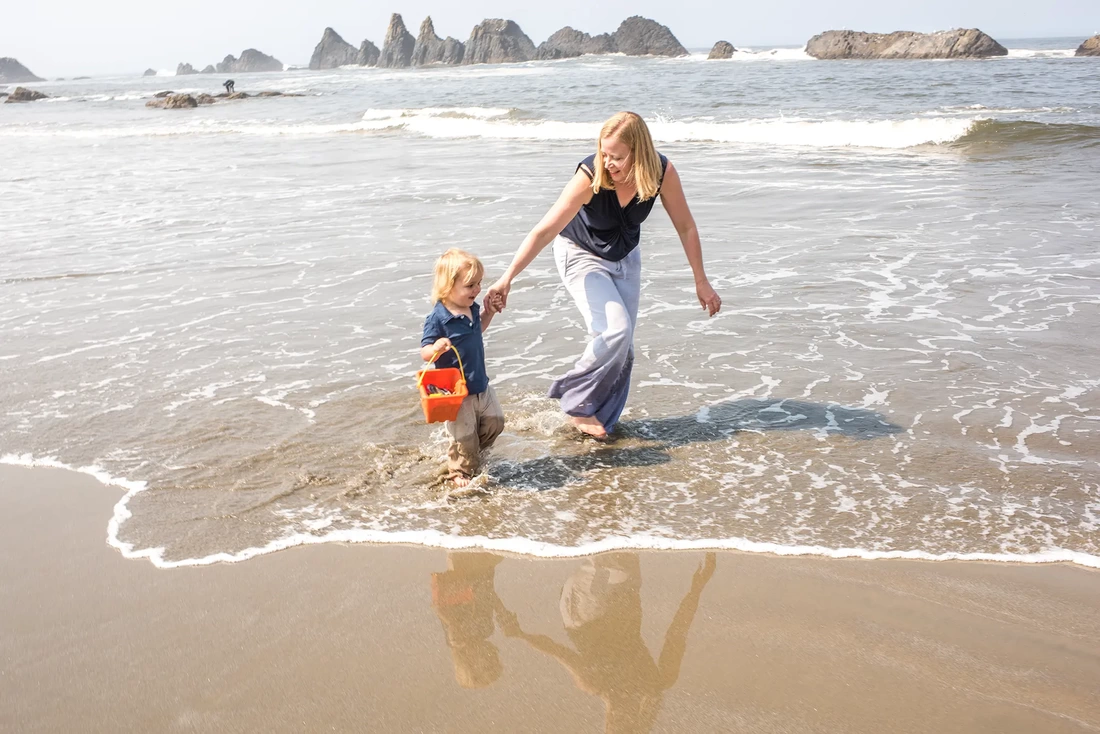A little boy and mother walk through the shallow waves together. They both smile. Rocks rise from the waves in the distance   Family Pictures Beach Theme with Portland Family Photographer Robert Knapp