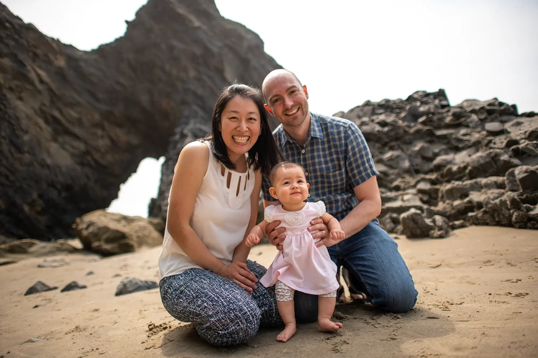 A family of three together on the sand. The mother and father smile brightly toward the camera. Baby smiles to the distance. Rocky features make interesting shapes in the background