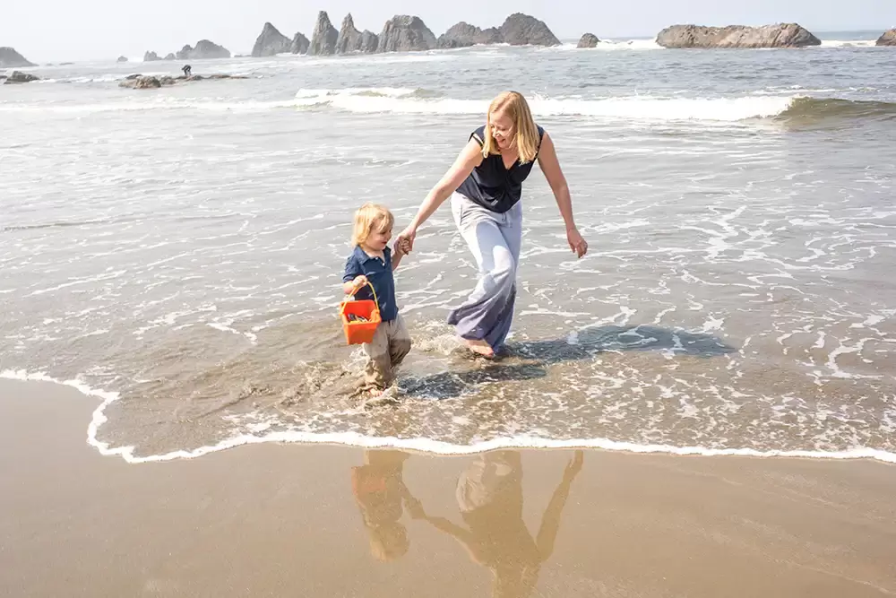 little boy smiles a big smile as the waves splash around his ankles. His mother leads him on.   Family Pictures Beach Theme with Portland Family Photographer Robert Knapp