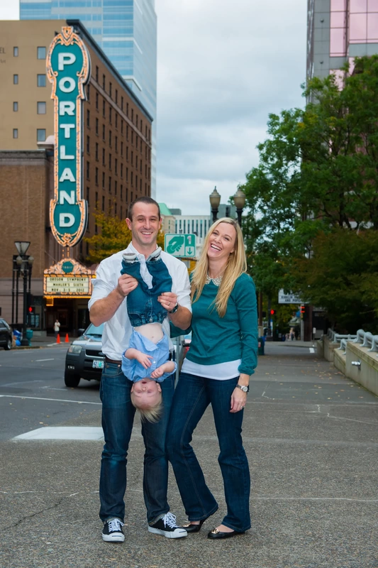 A father holds his son upsidedown in front of a marquee sign that reads PORTLAND, Mother holds the father and laughs. Family Photography by Robert Knapp in Portland City Parks