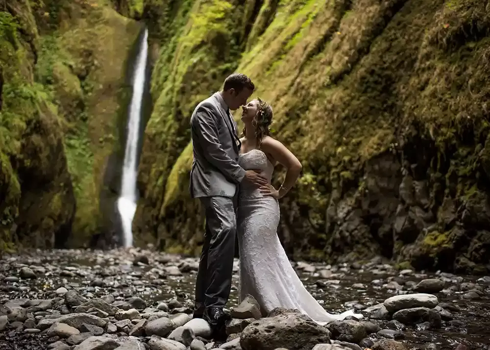 Photographers Portland at Trash the Dress Under a Waterfall. A couple stands on round river rocks as a waterfall stands behind them. The light is low in the ravine. The light is dark in the shadows. The couple look ready to kiss. Light green grass and moss contract the dark black shadows of the hills around. 